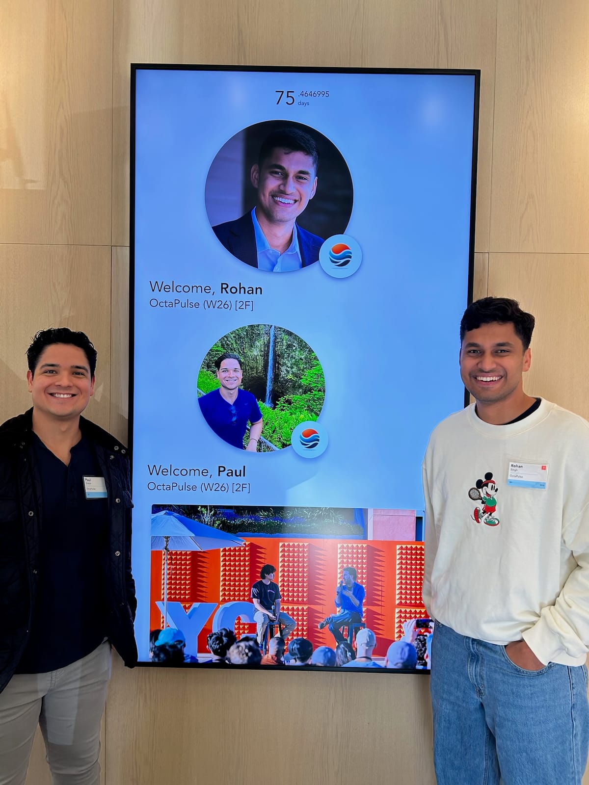 Rohan and Paul standing beside a YC lobby check-in screen showing both of their headshots, each captioned "OctaPulse (W26)", with a counter at the top reading 75.4646995 days