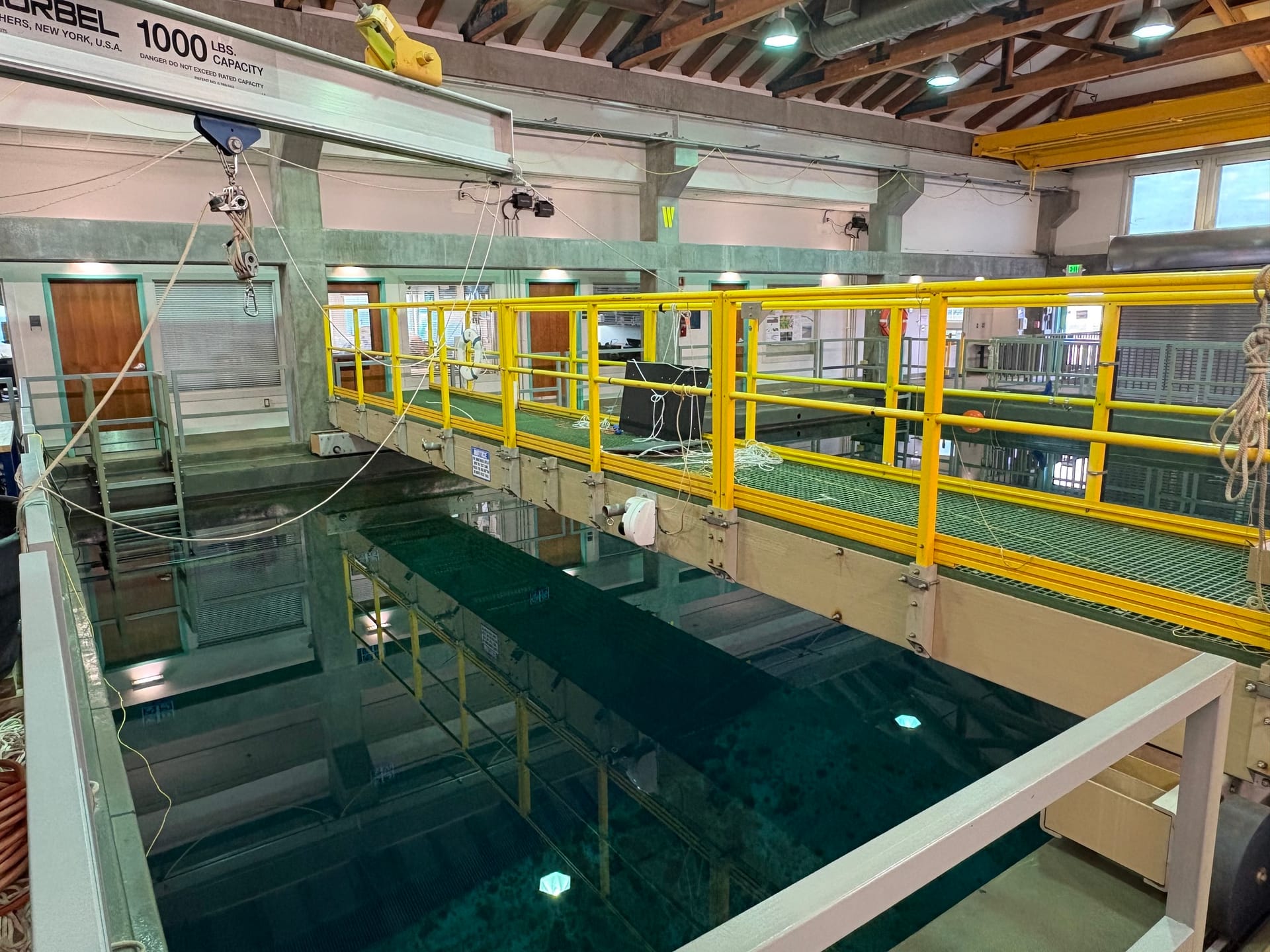 An indoor engineering test basin: a green-grating walkway with yellow tubular railing running above deep green water, an overhead Gorbel crane rail stamped 1000 LBS CAPACITY, cables and rope hanging down into the tank, timber roof trusses above