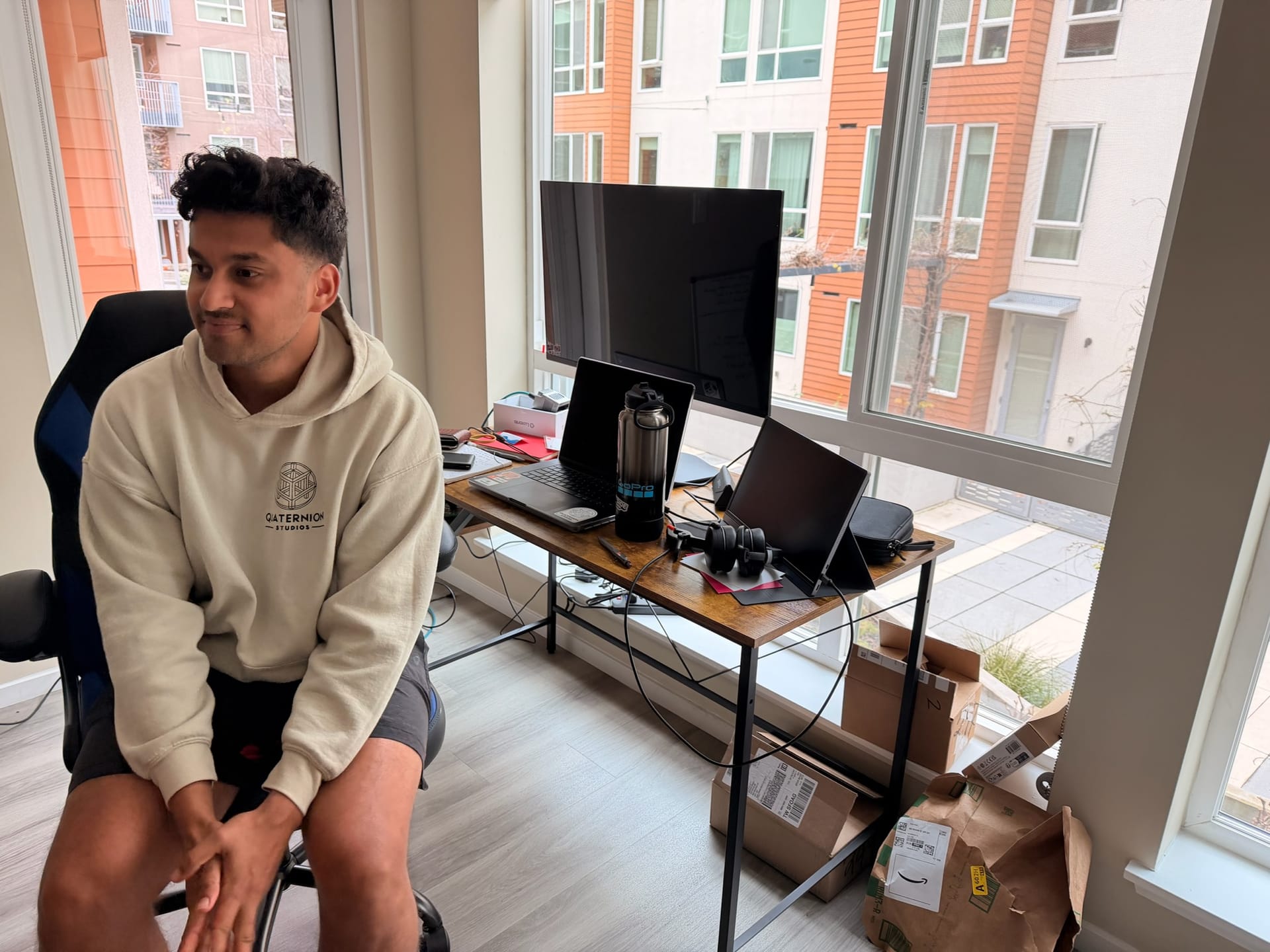 Rohan sitting in a black office chair beside a wood-top desk on a black metal frame, holding a laptop, an external monitor, a portable screen and headphones, with Amazon boxes on the floor and an apartment building through the windows