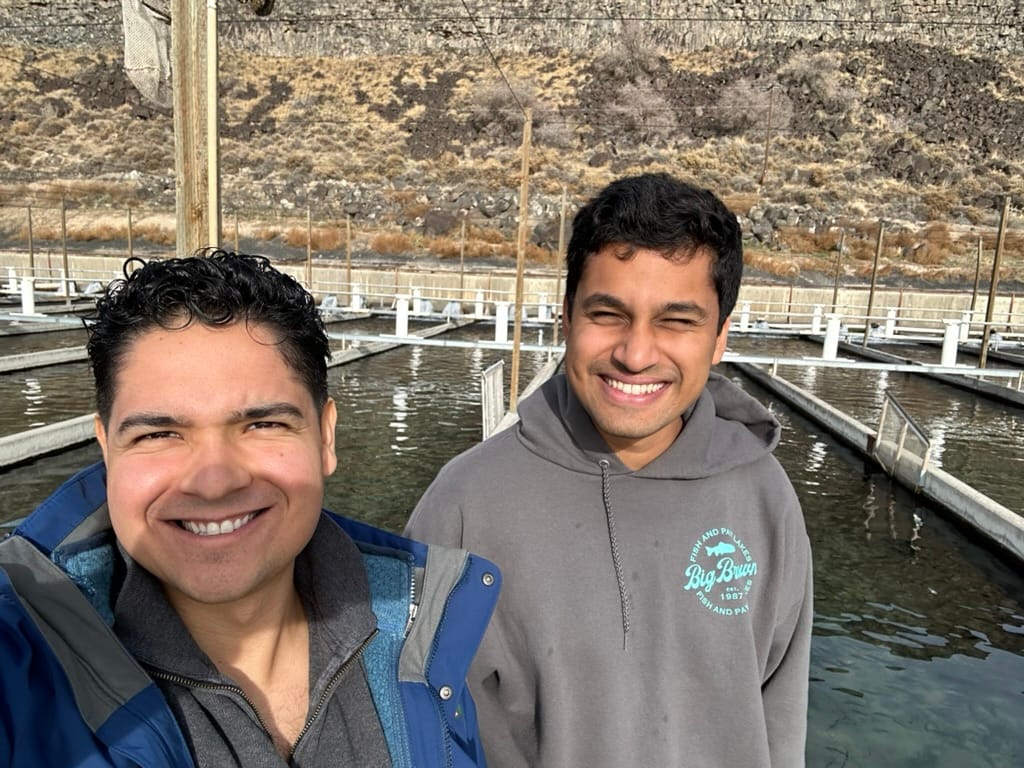 Rohan Singh and co-founder Paul Grech standing in front of long parallel concrete raceways at a trout farm, arid basalt hillside behind them