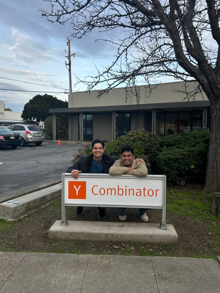 Two founders leaning together over a low white Y Combinator sign with the orange Y logo, a bare tree and parked cars behind them under an overcast sky