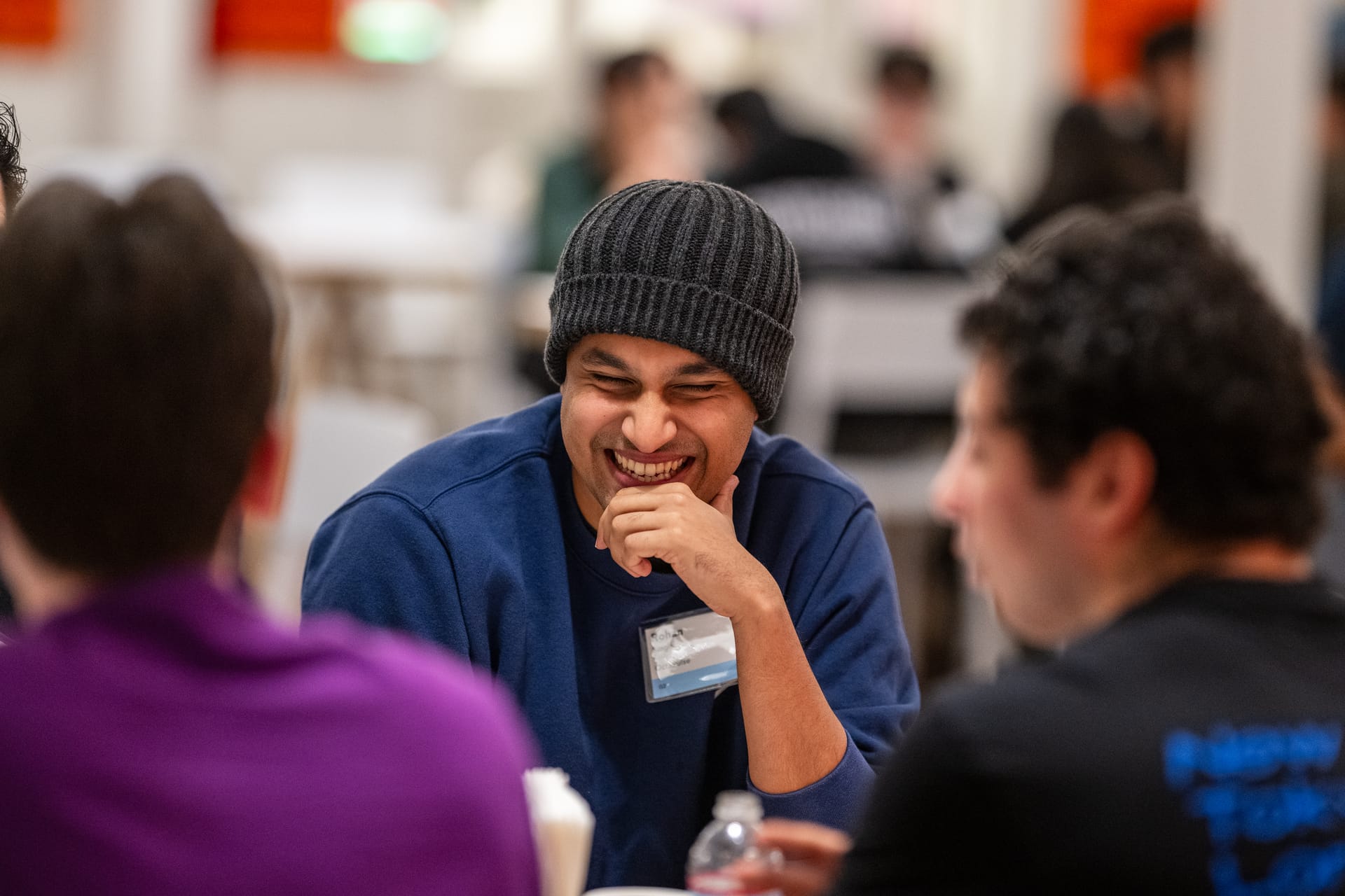 Rohan in a grey beanie and blue sweatshirt, laughing at a table with other founders at a YC event, a name badge on his chest