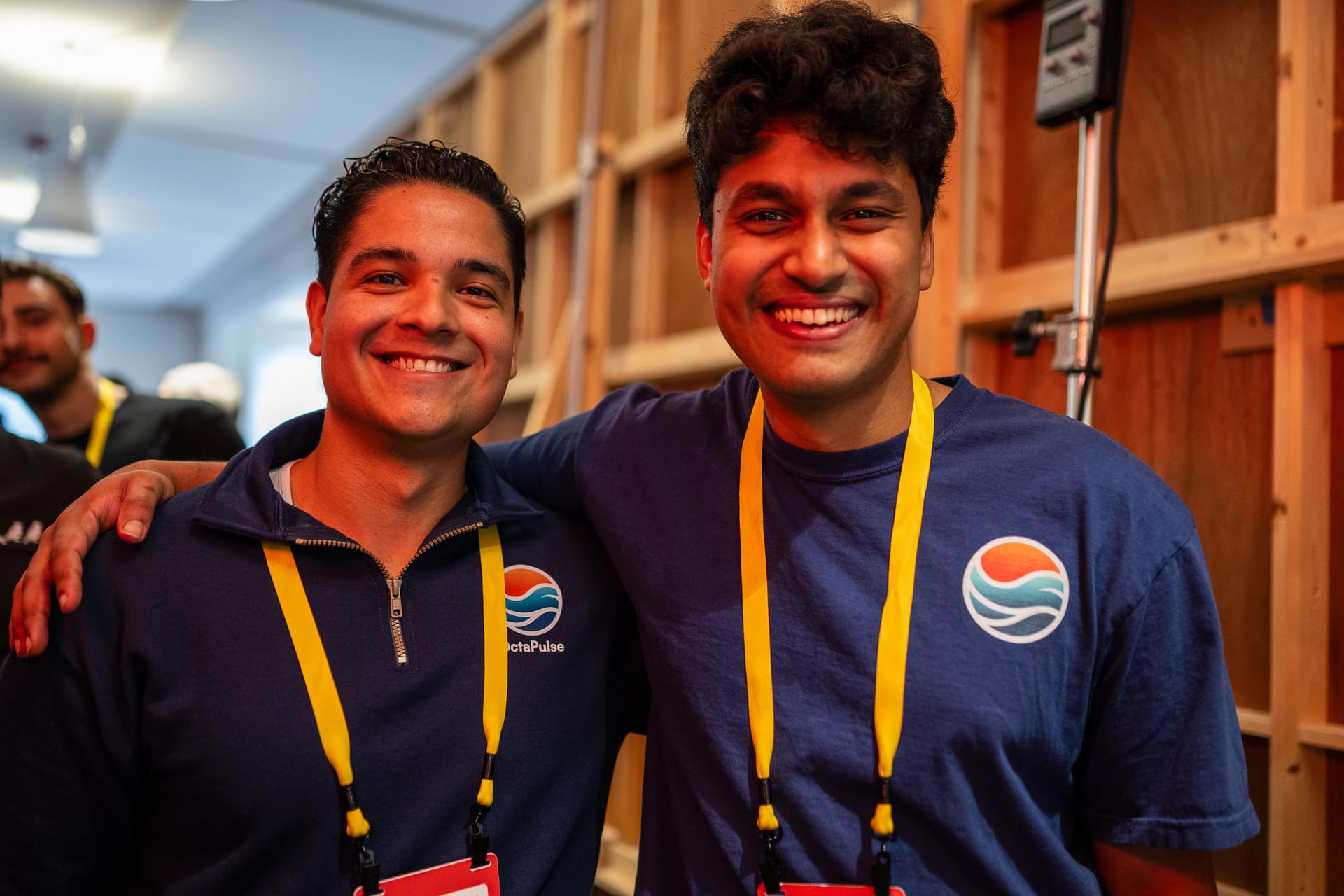 Paul Grech and me in navy OctaPulse-branded tops with yellow event lanyards, arms around each other's shoulders, standing in front of plywood shelving under warm indoor light