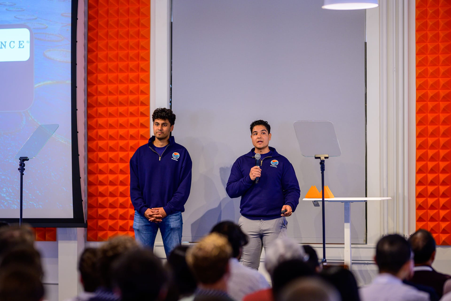 Rohan and Paul on stage in navy OctaPulse quarter-zips presenting to a seated audience, Paul on the microphone, orange acoustic paneling flanking the stage and a partner slide displayed behind them