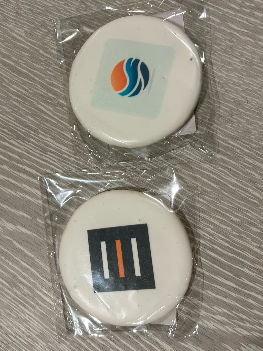 Two round iced cookies in clear cellophane on a grey wood-grain table: the top one printed with the OctaPulse logo, an orange and blue wave in a circle on a pale teal square, and the bottom one with the Pioneer Fund logo, three vertical bars on a dark square